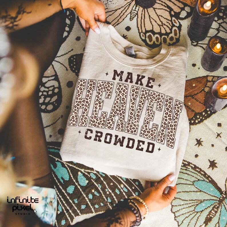 May include: A beige sweatshirt with the text "MAKE HEAVEN CROWDED" in a brown leopard print design. The sweatshirt is folded and held by hands, with a patterned cloth in the background.