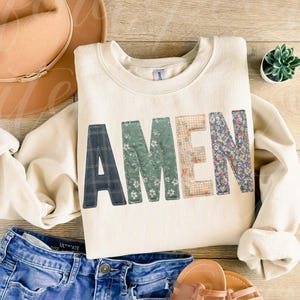 May include: Cream-colored sweatshirt with the word "AMEN" in patchwork letters. The letters are made of various patterned fabrics, including plaid, floral, and gingham. The sweatshirt is paired with blue jeans, sandals, and a hat.