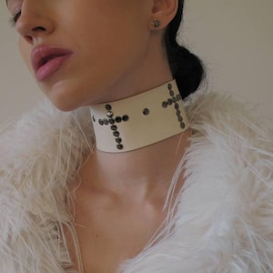 May include: A cream-colored choker with a cross design made of small, dark studs. The choker is worn over a white, fluffy faux fur coat. The image is a close-up of the neck and upper chest.