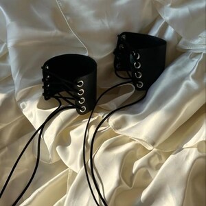 May include: Two black leather wrist cuffs with silver eyelets and black lacing. The cuffs are cylindrical and rest on a cream-colored, silky fabric. The lacing extends from the cuffs, creating a decorative element.