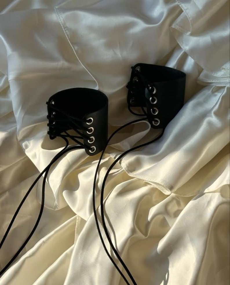 May include: Two black leather wrist cuffs with silver eyelets and black lacing. The cuffs are cylindrical and rest on a cream-colored, silky fabric. The lacing extends from the cuffs, creating a decorative element.