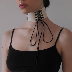 May include: Cream-colored choker necklace with black lacing and ties. The choker is made of a thick material and has a lace-up design in the front. The black ties hang down from the choker.