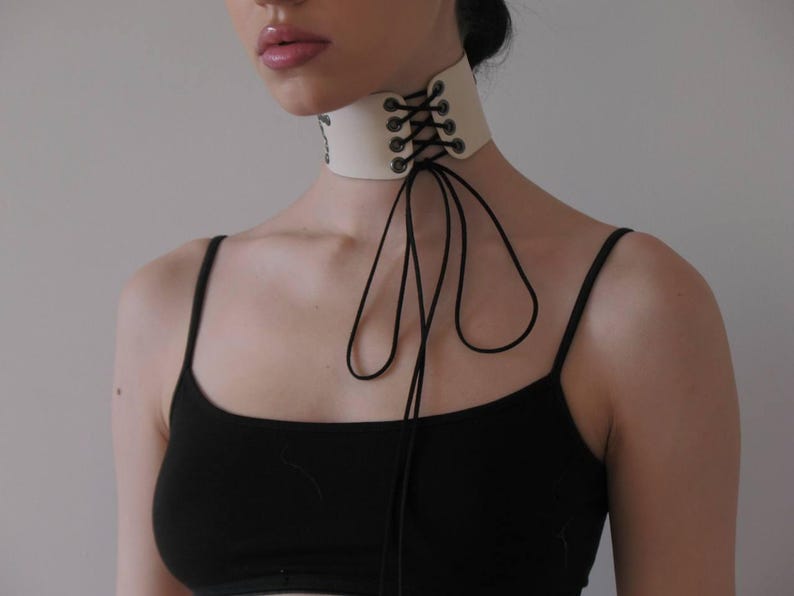 May include: Cream-colored choker necklace with black lacing and ties. The choker is made of a thick material and has a lace-up design in the front. The black ties hang down from the choker.