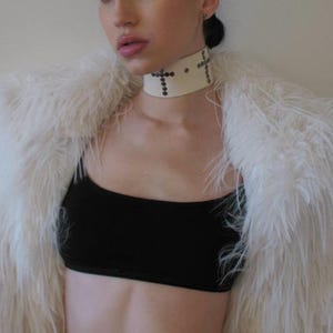 May include: A cream-colored choker with cross-shaped embellishments and small studs. The choker is worn with a black top and a fluffy white faux fur coat.