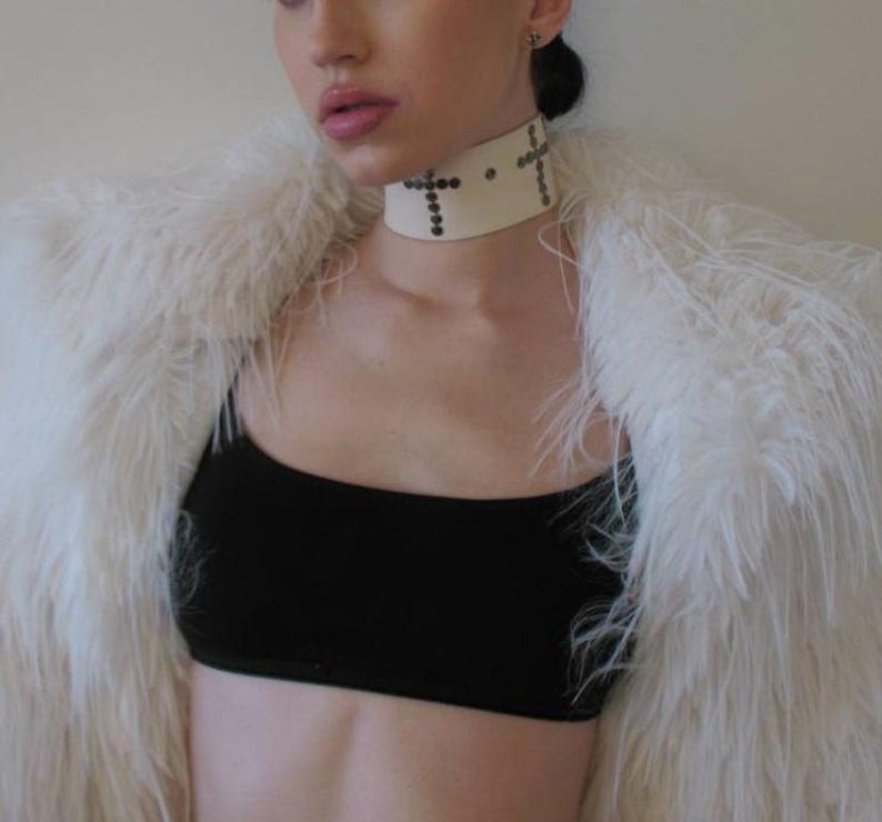 May include: A cream-colored choker with cross-shaped embellishments and small studs. The choker is worn with a black top and a fluffy white faux fur coat.