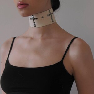 May include: Cream-colored choker necklace with silver-toned cross designs. The wide collar is adorned with small, round studs. The model is wearing a black tank top.
