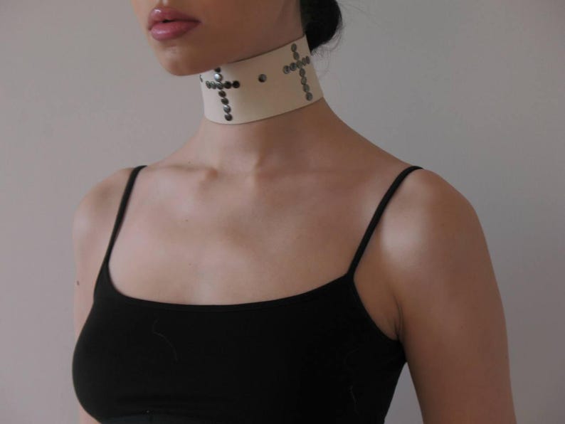 May include: Cream-colored choker necklace with silver-toned cross designs. The wide collar is adorned with small, round studs. The model is wearing a black tank top.