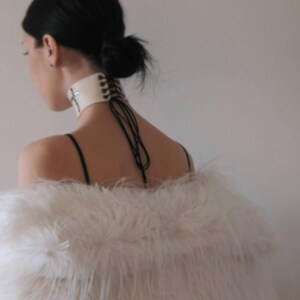 May include: A cream-colored choker with a cross design and black lacing detail. The choker is worn with a white feathered shawl. The model has dark hair pulled back in a bun.