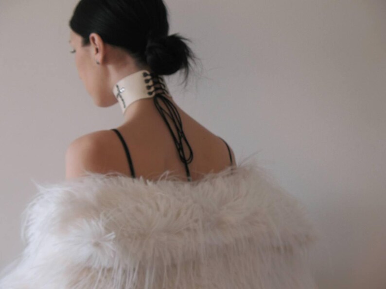May include: A cream-colored choker with a cross design and black lacing detail. The choker is worn with a white feathered shawl. The model has dark hair pulled back in a bun.