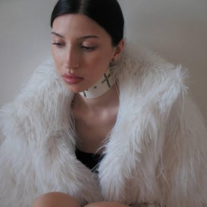 May include: A white faux fur coat with a wide collar. The model is wearing a white choker necklace with cross details. The person is wearing a black top.