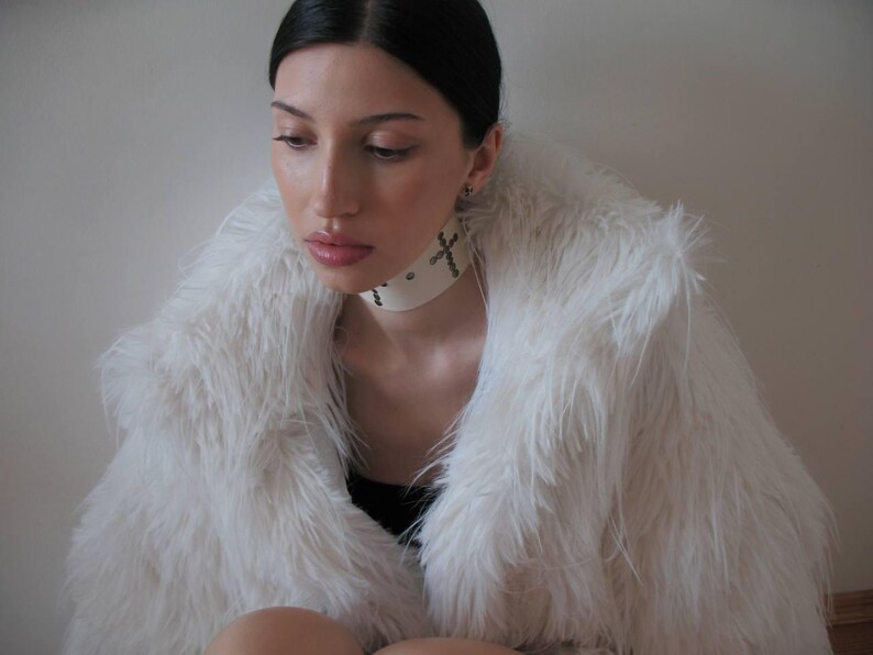 May include: A white faux fur coat with a wide collar. The model is wearing a white choker necklace with cross details. The person is wearing a black top.