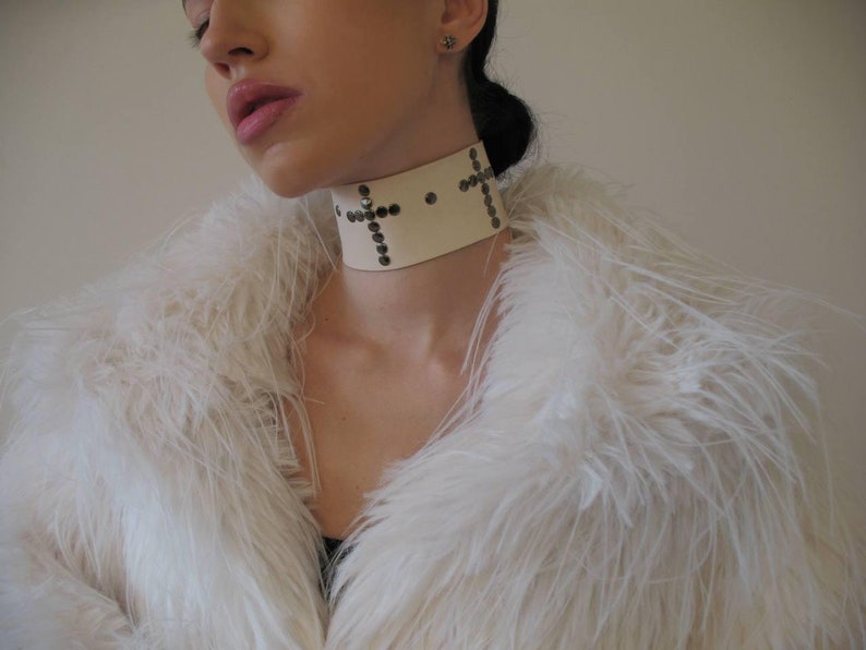 May include: A cream-colored choker with a cross design made of small, dark studs. The choker is worn over a white, fluffy faux fur coat. The image is a close-up of the neck and upper chest.