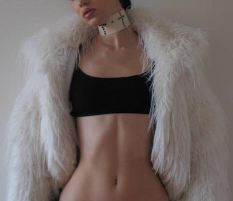 May include: A white faux fur coat draped over a black crop top. A cream-colored choker with black text is worn around the neck. The coat has long, fluffy fibers.