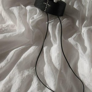 May include: A black leather choker with a silver cross design and small silver studs. The choker has two long black cords hanging down. The choker is displayed on a white, textured fabric.