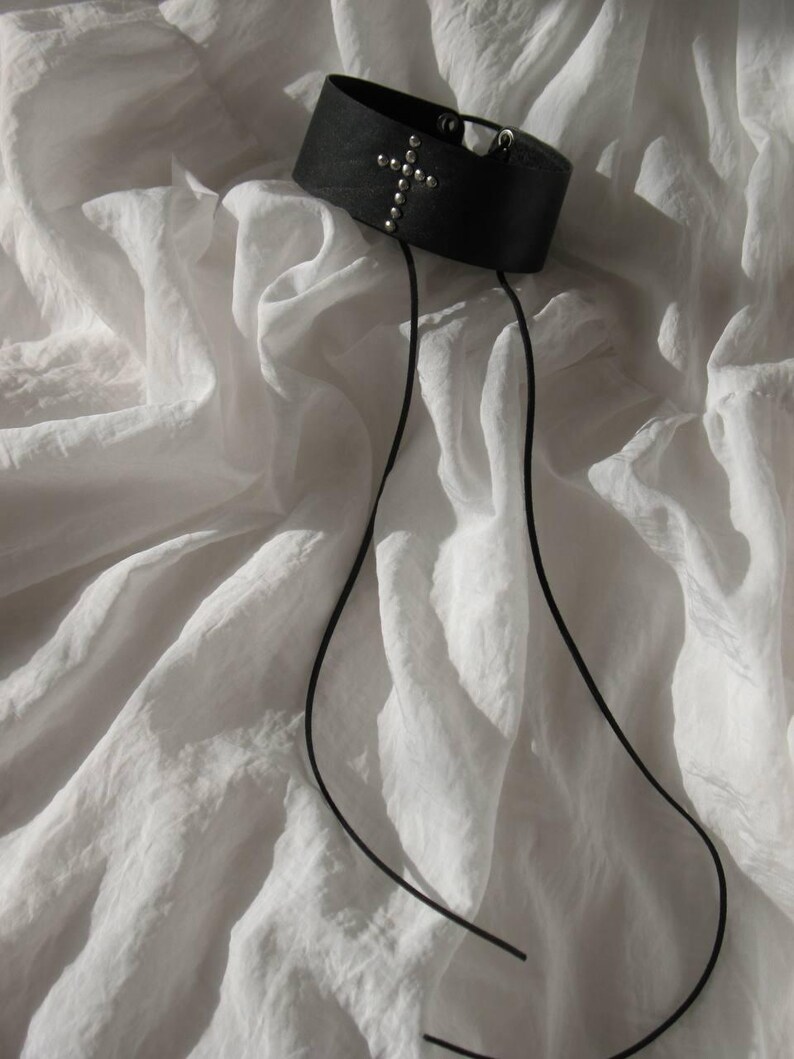 May include: A black leather choker with a silver cross design and small silver studs. The choker has two long black cords hanging down. The choker is displayed on a white, textured fabric.