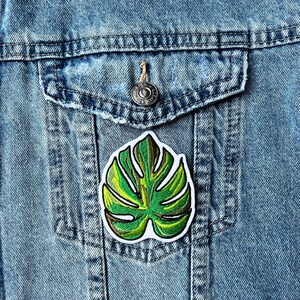 May include: A green and yellow embroidered monstera leaf patch with a white border is attached to the pocket of a blue denim jacket. The jacket has a button closure and a pocket flap.
