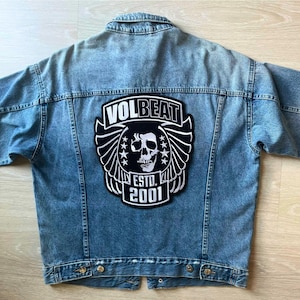 May include: Light blue denim jacket with a large black and white patch on the back. The patch features the band name "VOLBEAT", a skull, wings, stars, and the text "ESTD. 2001". The jacket has a classic button-up design.