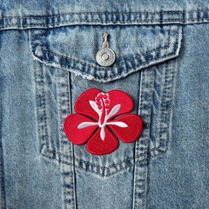 May include: A red hibiscus flower patch with white detailing is sewn onto the pocket of a blue denim jacket. The patch is embroidered with detailed stitching. The jacket has a button closure and a classic design.