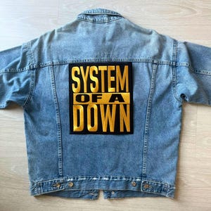 Puede incluir: Chaqueta vaquera azul claro con un gran parche negro en la espalda. El parche presenta las palabras "SYSTEM OF A DOWN" en letras doradas. La chaqueta tiene un diseño clásico con botones y bolsillos.