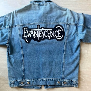 May include: A light blue denim jacket with a large black and white patch on the back. The patch displays the band name "EVANESCENCE" in a stylized font. The jacket has a classic design with button closures and pockets.