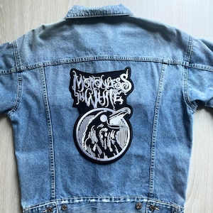 Pode incluir: Jaqueta jeans azul claro com um grande patch com o nome da banda "Motionless in White" e uma ilustração de corvo em preto e branco. A jaqueta tem um design clássico com fechos de botão e bolsos.