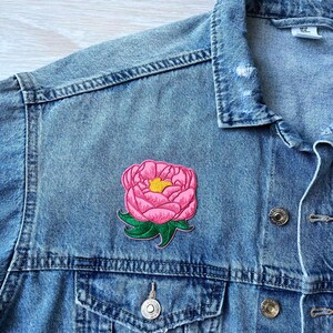 Pode incluir: Um casaco de ganga azul claro com um emblema de peónia cor-de-rosa com centro amarelo e folhas verdes. O casaco tem um colarinho clássico, botões de metal e um bolso no peito. O tecido apresenta uma trama visível e um acabamento lavado.