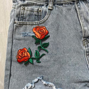 Puede incluir: Shorts vaqueros azul claro con dos rosas naranjas bordadas y hojas verdes. Los shorts tienen un aspecto desgastado con una sección rasgada. Diseño clásico de cinco bolsillos.