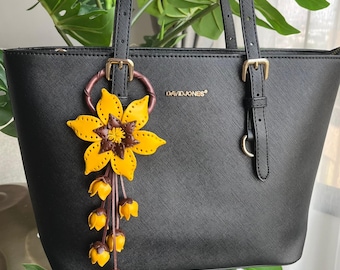 Könnte beinhalten: Eine schwarze Ledertasche mit einem goldenen Reißverschluss und einem Schlüsselanhänger aus Leder in Form einer Sonnenblume. Der Schlüsselanhänger hat eine braune Mitte und gelbe Blütenblätter. Die Tasche trägt den Schriftzug "DAVID JONES" auf der Vorderseite.