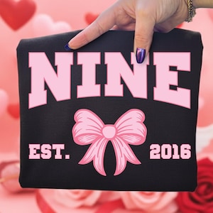 Puede incluir: Prenda negra con la palabra "NINE" en rosa, un lazo rosa y el texto "EST. 2016". La prenda es sostenida por una mano con uñas pintadas, sobre un fondo rosa borroso.