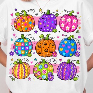 May include: A white t-shirt featuring a graphic print of nine colorful pumpkins arranged in three rows. The pumpkins display various patterns including polka dots, stars, flowers, hearts, jack-o'-lantern faces, and swirls, accented by small stars and hearts.