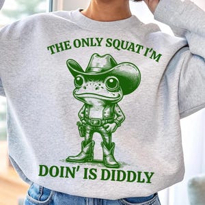Może przedstawiać: Szara bluza z zieloną żabą w kowbojskim kapeluszu i napisem "The only squat I'm doin' is diddly".