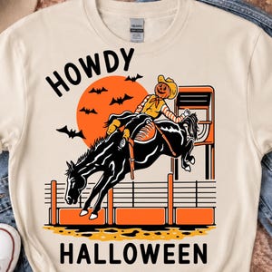 May include: A cream-colored t-shirt featuring a graphic of a pumpkin-headed figure in a cowboy hat riding a bucking black horse. The design includes a large orange moon, black bats, a rodeo chute, and the text HOWDY HALLOWEEN in black letters.