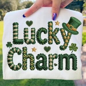 Puede incluir: Tela blanca con las palabras "Lucky Charm" bordadas en hilo verde y dorado. El diseño incluye un sombrero de duende verde, tréboles, corazones y estrellas, ideal para el Día de San Patricio.