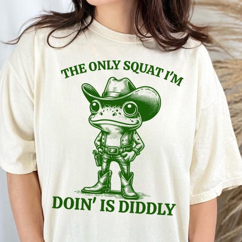 Może przedstawiać: Biały t-shirt z zieloną ilustracją żaby w kowbojskim kapeluszu i tekstem "The only squat I'm doin' is diddly."