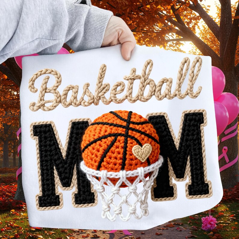 May include: White fabric with the words "Basketball Mom" in a rope-like font. The "MOM" is in black with a white border. A crocheted basketball with a heart is in the center.