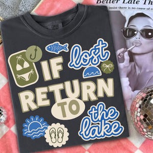 Könnte beinhalten: Dunkelgraues T-Shirt mit dem Text "If lost return to the lake" in verspielter Schrift. Das Shirt zeigt verschiedene Grafiken zum Thema See, darunter ein Fisch, Bikini und Flip-Flops. Ein Comfort Colors Markenetikett ist sichtbar.