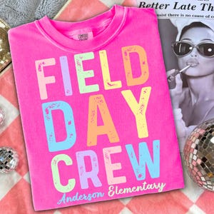 Puede incluir: Una camiseta rosa brillante con las palabras "FIELD DAY CREW" en letras grandes y coloridas. El texto "Anderson Elementary" está impreso debajo. La camiseta es de material suave y tiene cuello redondo.
