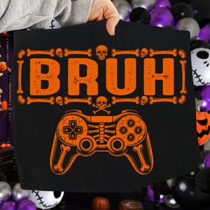May include: Black t-shirt with an orange graphic. The graphic features the word "BRUH" in a bone-like font, skulls, and a skeleton-themed video game controller. A Halloween-themed design.