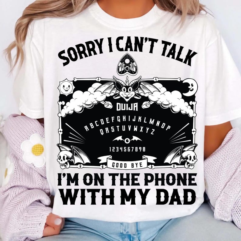 May include: White t-shirt with a black Ouija board graphic. The design includes the alphabet, numbers, and the words "Sorry I Can't Talk, I'm on the Phone with My Dad."