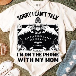 May include: White t-shirt with a black Ouija board graphic. The design includes the text "SORRY I CAN'T TALK I'M ON THE PHONE WITH MY MOM". The graphic features a bat, skulls, and the alphabet.