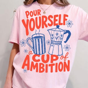 May include: A light pink t-shirt featuring a graphic print in red and blue. The design includes a striped mug, a moka pot, and small flower icons, with the text "POUR YOURSELF a CUP of AMBITION" written in a bold, stylized red font across the front.