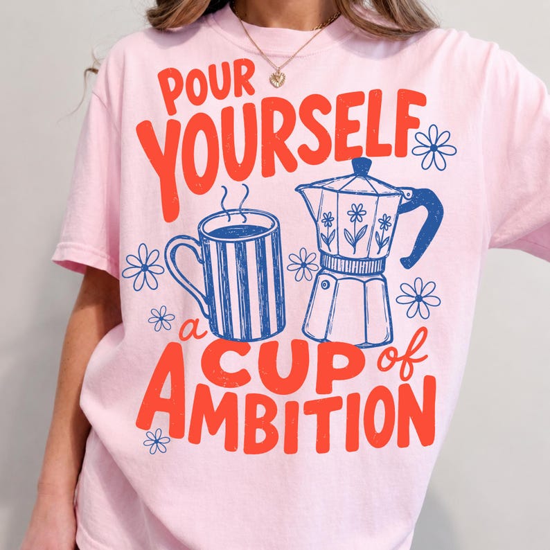 May include: A light pink t-shirt featuring a graphic print in red and blue. The design includes a striped mug, a moka pot, and small flower icons, with the text "POUR YOURSELF a CUP of AMBITION" written in a bold, stylized red font across the front.