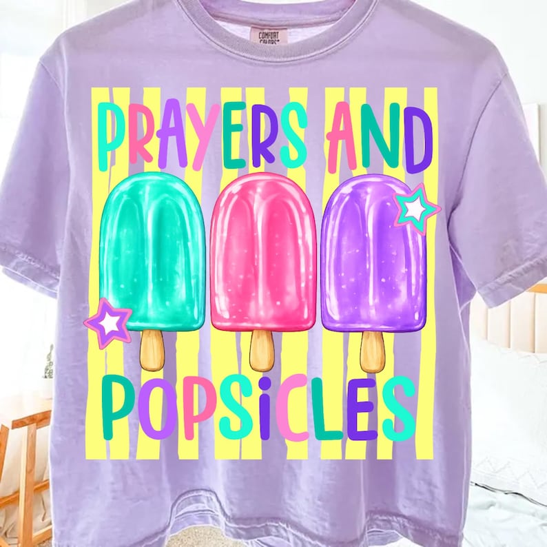 K&ouml;nnte beinhalten: Helllila T-Shirt mit der Aufschrift "PRAYERS AND POPSICLES" in bunten Farben. Das Design zeigt drei Eis am Stiel-Illustrationen in T&uuml;rkis, Rosa und Lila, mit gelben vertikalen Streifen im Hintergrund.