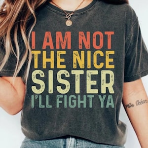 Puede incluir: Camiseta gris oscuro con el texto "I AM NOT THE NICE SISTER I'LL FIGHT YA" en letras naranjas, amarillas y verdes. La camiseta tiene un aspecto vintage y desgastado.