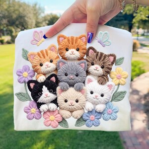 Pu&ograve; includere: Un oggetto di tessuto bianco con sette gattini ricamati in vari colori, circondati da fiori e farfalle colorate. I gattini includono disegni arancioni, grigi, marroni, bianchi e neri e color crema.
