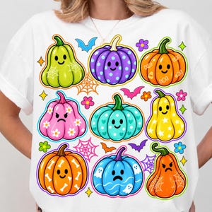 May include: A white t-shirt featuring a colorful graphic print of nine stylized pumpkins with various facial expressions and patterns, including polka dots, stars, bones, and spiderwebs, interspersed with small bats, flowers, and spiderweb icons.