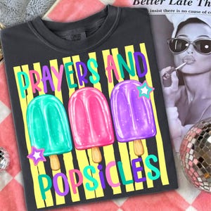 K&ouml;nnte beinhalten: Dunkelgraues T-Shirt mit den Worten "Prayers and Popsicles" in farbenfrohem Text. Das Shirt zeigt drei Eis am Stiel-Illustrationen in Gr&uuml;n, Pink und Lila, jeweils mit einem Stern. Marke Comfort Colors.