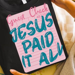May include: Black t-shirt with a pink graphic resembling a guest check. The graphic reads "Guest Check" in pink script, with the words "JESUS PAID IT ALL" in teal, and a number "482759".