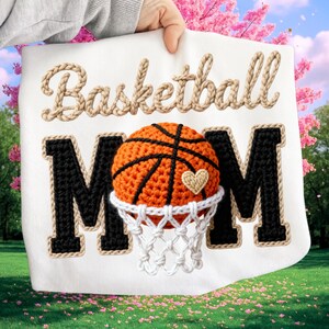 May include: White fabric with the words "Basketball MOM" in a decorative font. The word "Basketball" is in a rope-like font. The "MOM" is in a black block letter style with a rope-like border. A crocheted basketball is in the center, with a heart and net.