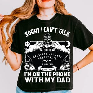 May include: Black t-shirt with a white graphic of a Ouija board, a bat, and the text "SORRY I CAN'T TALK" and "I'M ON THE PHONE WITH MY DAD". The design includes the alphabet, numbers, and the word "DUIJA".